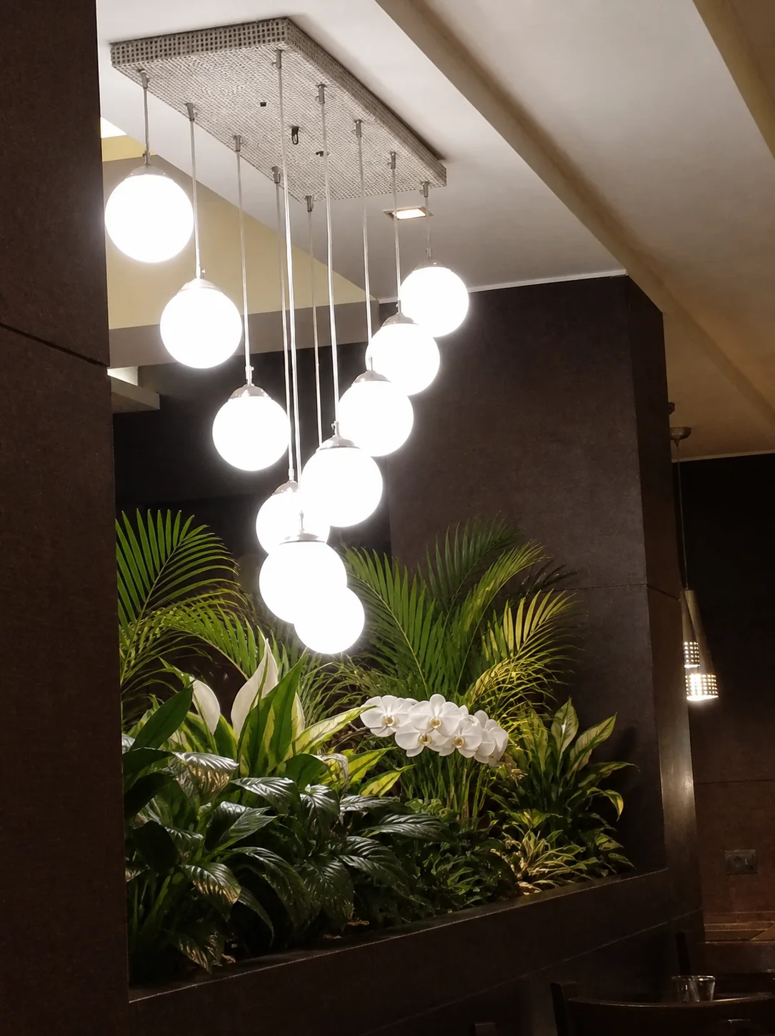Interior feature planter at Country Inn Gurugram with compact palms, broad foliage, white flowers and pendant lighting.