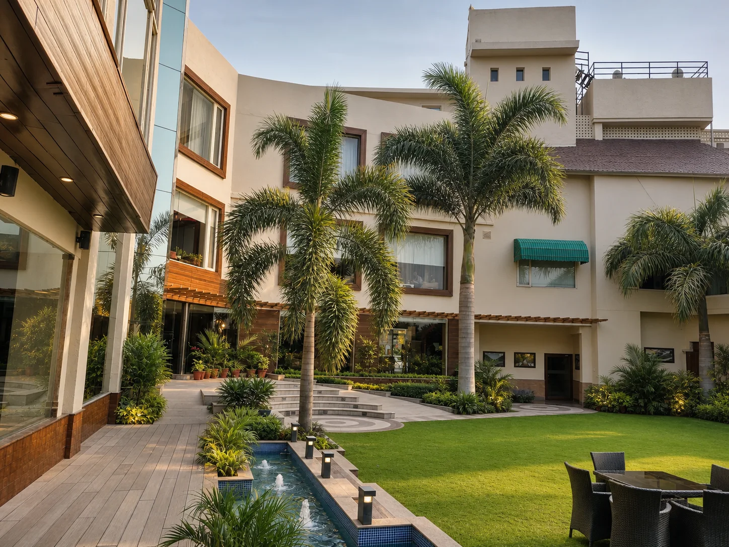 Country Inn Jammu courtyard with feather palms, lawn, layered tropical planting and a narrow fountain channel beside timber decking.