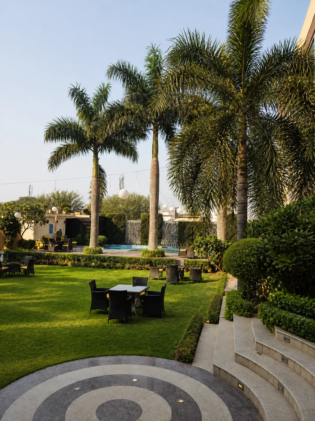Country Inn Jammu outdoor garden lounge with mature palms, lawn furniture, clipped borders and pool-garden views.