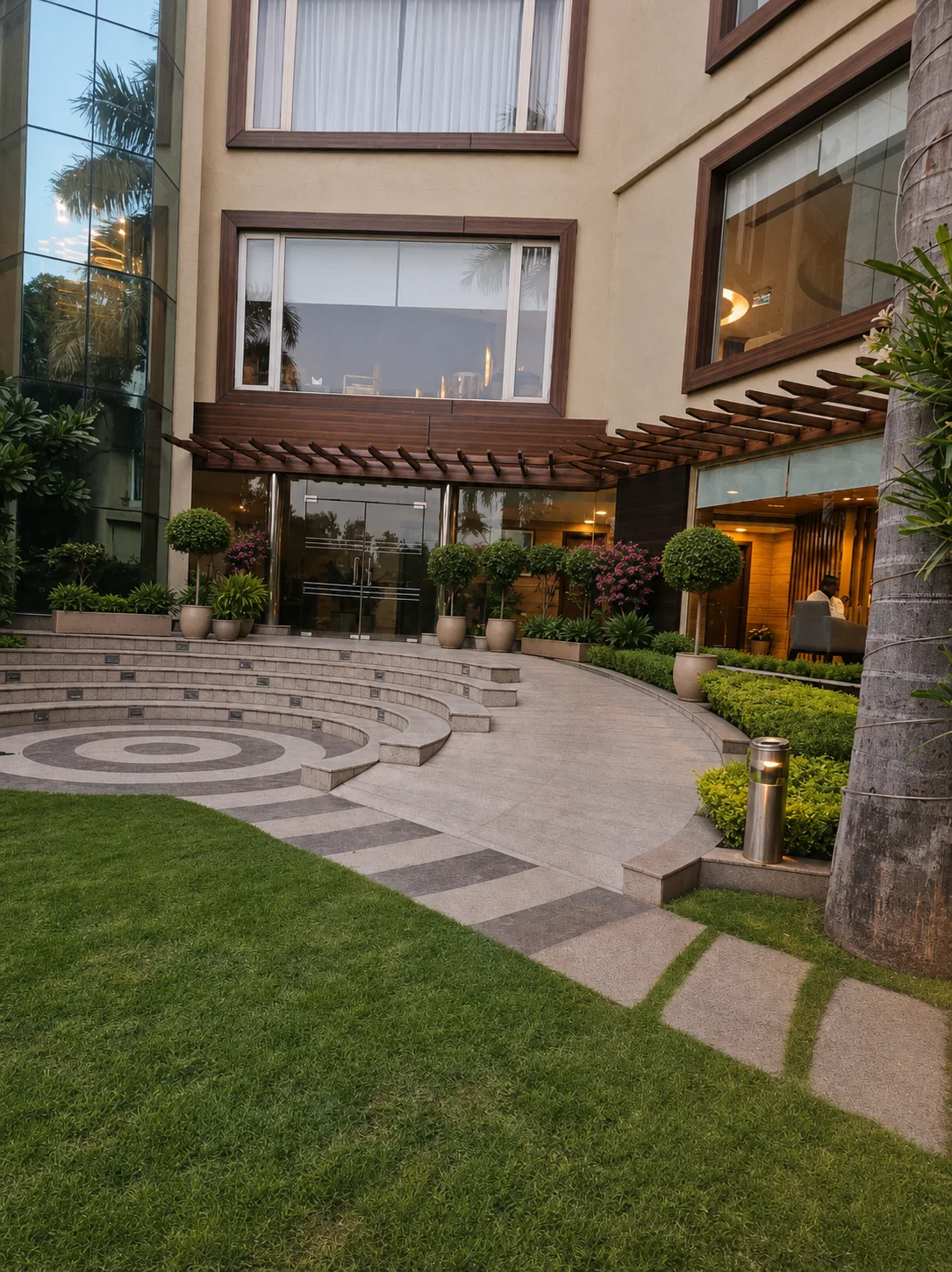 Country Inn Jammu garden lobby entrance with curved stone steps, timber pergola, topiary, tropical planting and warm interior light.