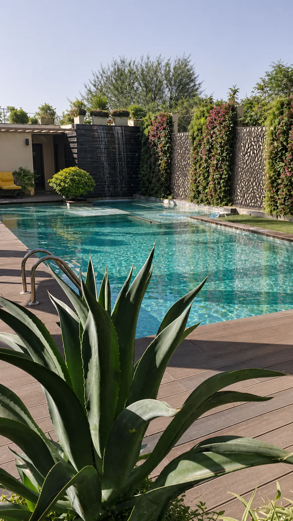 Country Inn Jammu pool view toward black-stone waterfalls, decorative screens and dense planted walls framed by an agave.