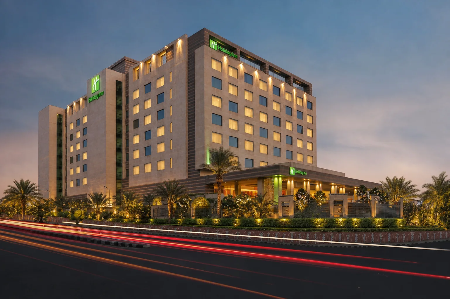 Holiday Inn Jaipur hotel facade framed by palms, evergreen hedges and white flowering accents at blue hour.