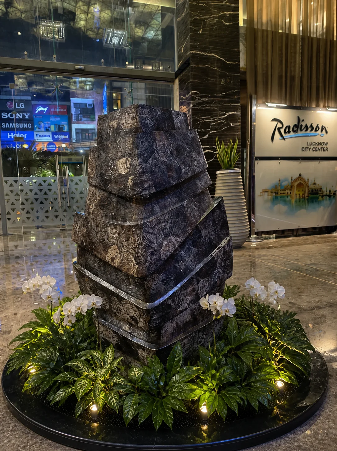 Radisson Lucknow lobby sculpture surrounded by glossy tropical foliage, white orchids and subtle pin lighting.