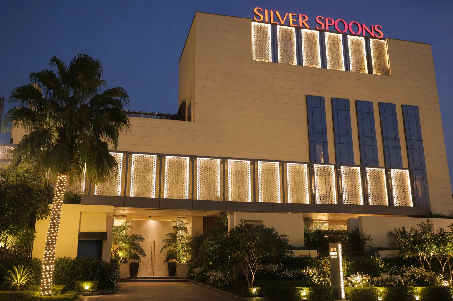 Silver Spoons Ghaziabad facade at blue hour with mature palms, layered evergreen planting and restrained white flowers.