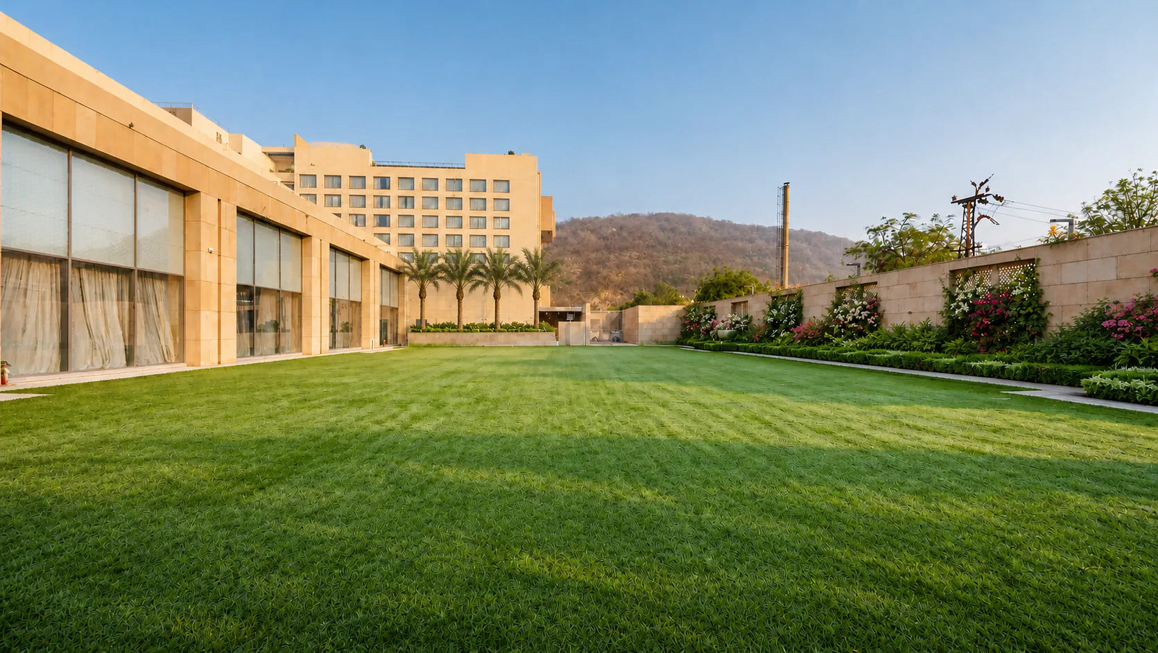 Taj Amer Jaipur event lawn in morning light with a broad turf area, formal perimeter planting and palms.