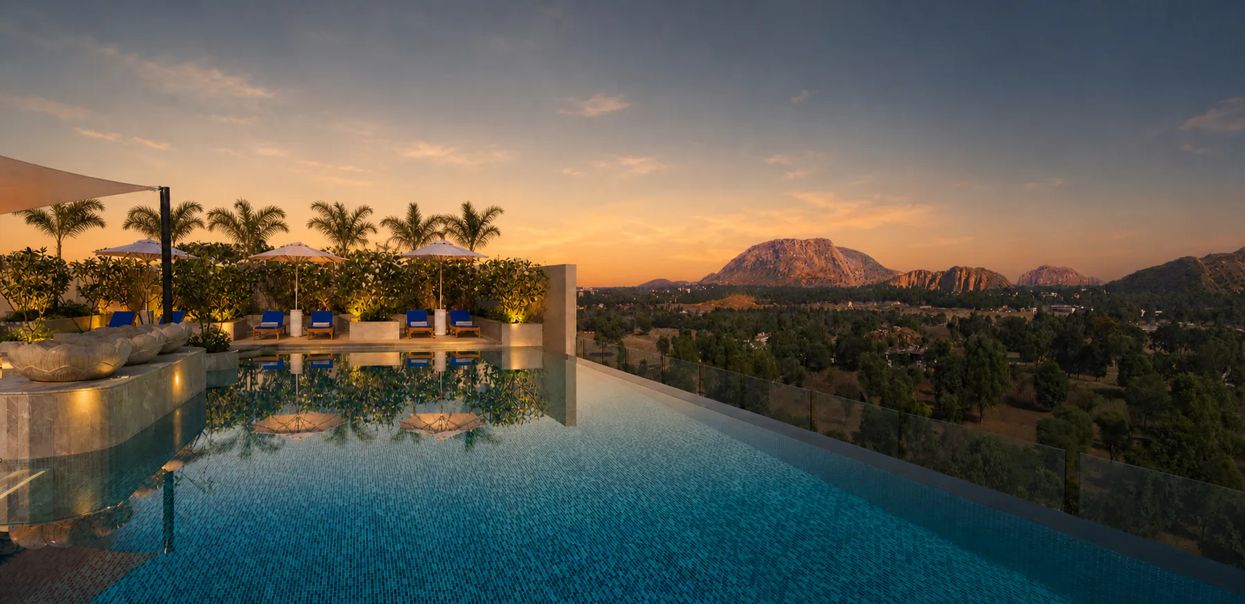 Taj Amer Jaipur infinity pool at golden hour with illuminated planters, foxtail palms and an Aravalli mountain view.