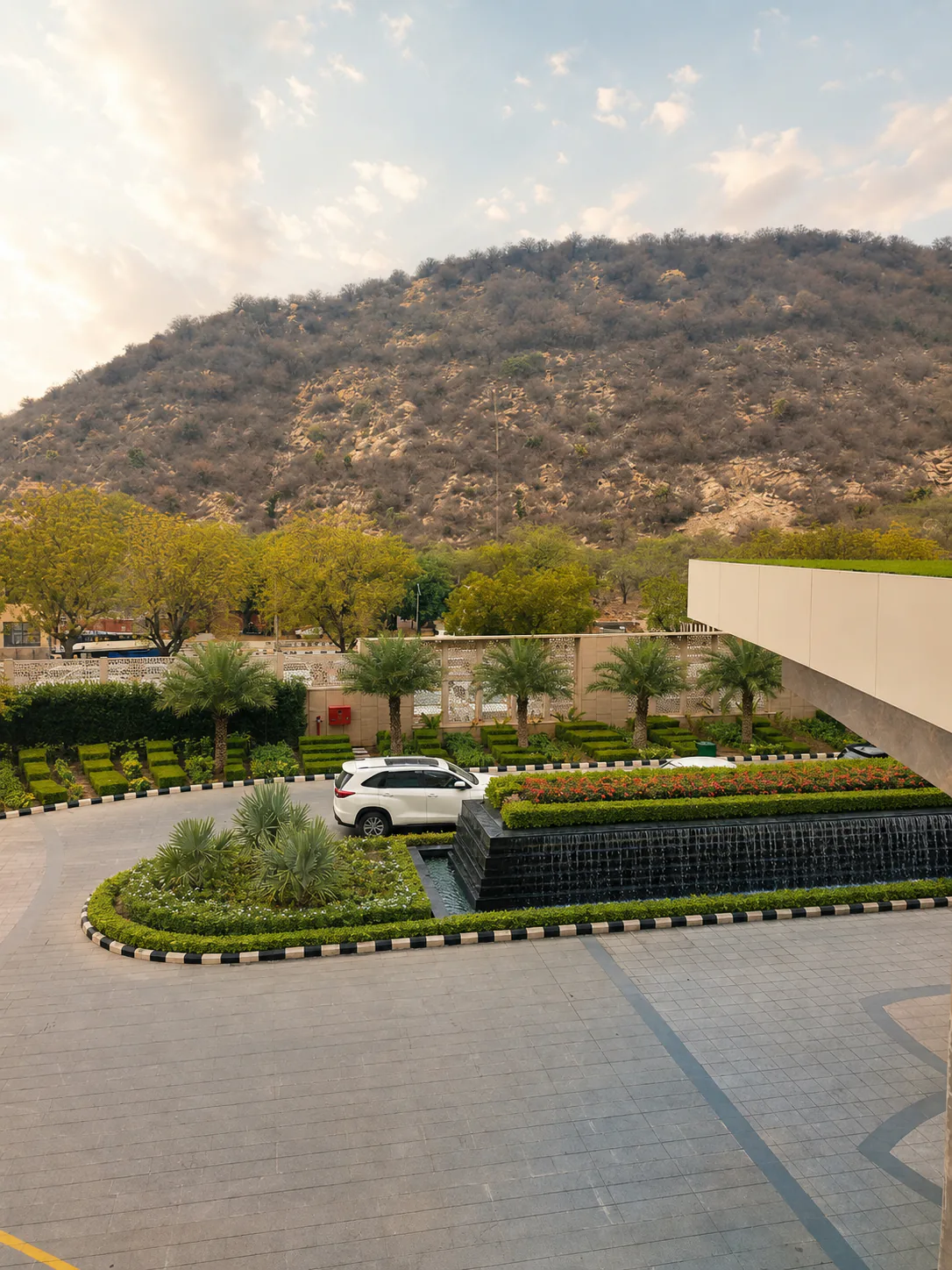 Elevated Taj Amer Jaipur arrival court with a black water wall, circular palm island and Aravalli backdrop.