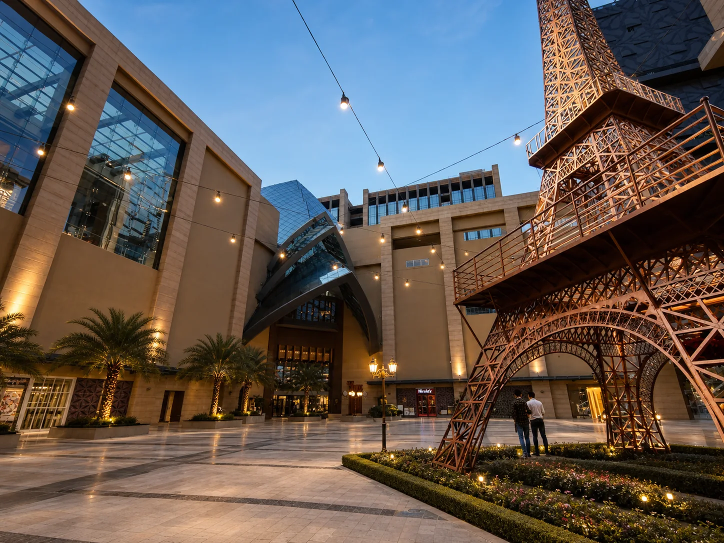 Vegas Mall Dwarka Eiffel-themed plaza at evening with planted tower base, entrance palms, string lights and open paving.