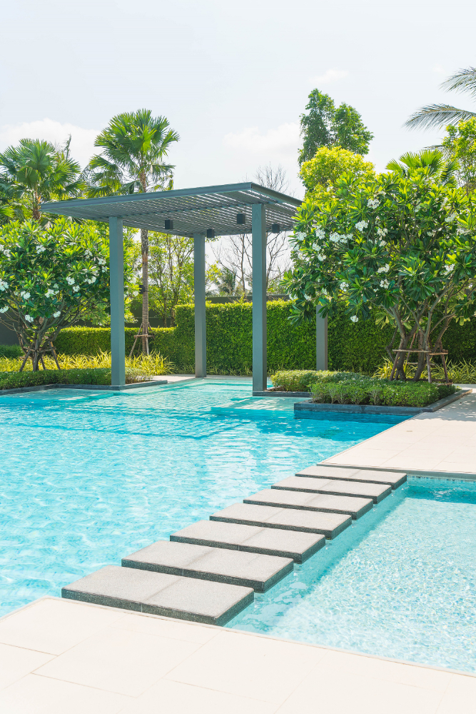 An elegant poolside landscape with a modern pergola and stepping stones leading across the shimmering blue water, surrounded by lush greenery, designed by Four Leaf Landscape.